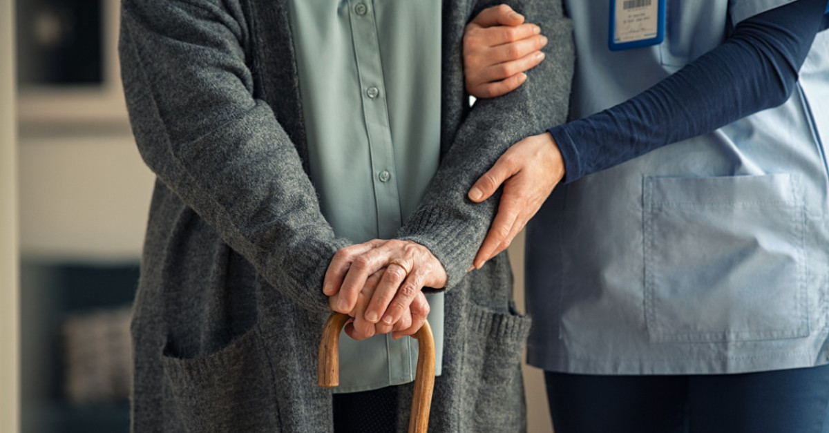 Older person walking with cane being assisted by nurse